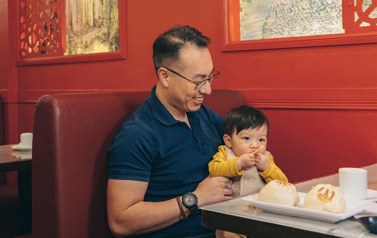 A man and a baby sit at a table with food, enjoying a meal together.